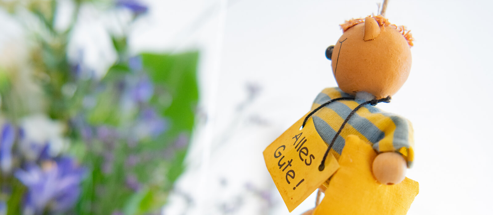 Foto mit Blumenstrauß und einer Holzfigur, die ein Schild mit der Aufschrift „Alles Gute!” trägt